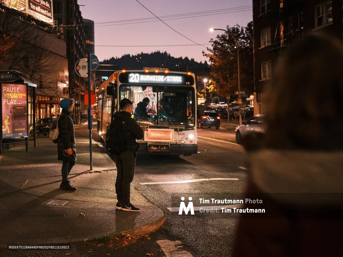 In the dusky twilight of Portland's Northwest District, passengers wait at a TriMet bus stop as the Route 20 Burnside/Stark line approaches. The amber glow of street lights mingles with the bus's bright headlights, casting warm pools of illumination across the wet asphalt of West Burnside Street. Two silhouetted figures in winter clothing stand patiently on the sidewalk, embodying the quiet rhythm of urban evening commutes against the backdrop of Portland's characteristic mixed-use architecture and forested hills.