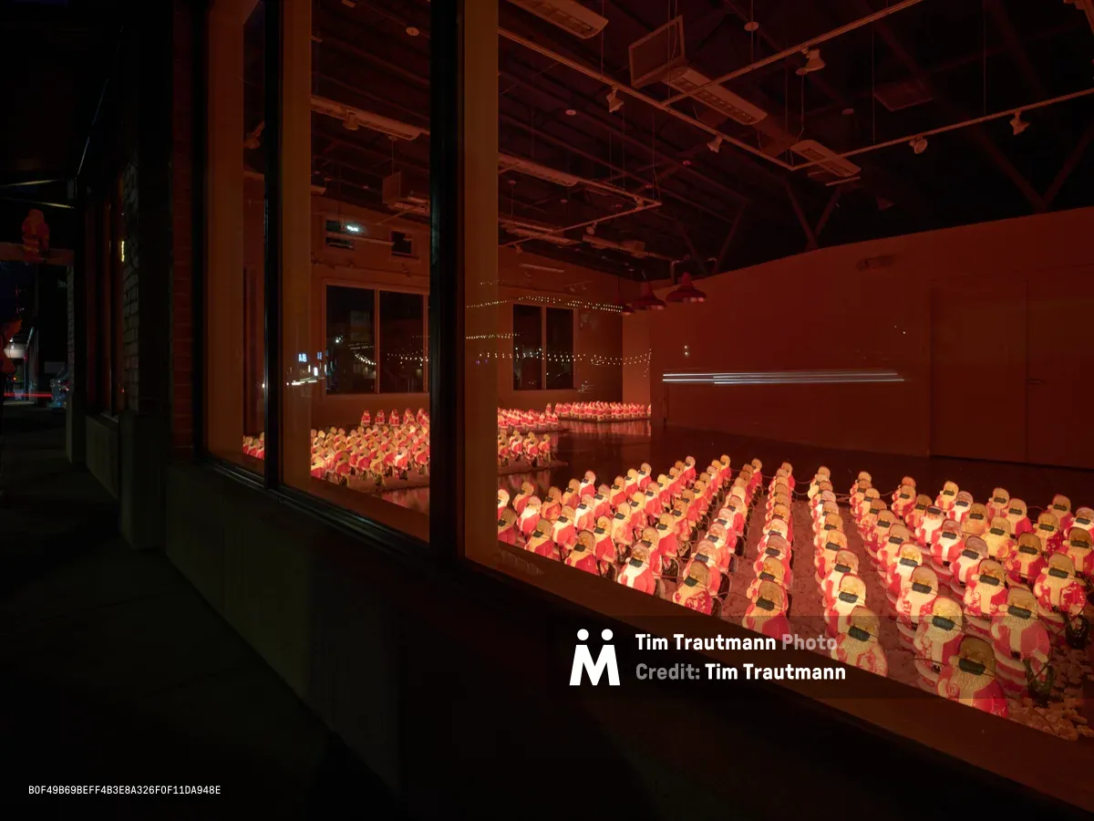 Hundreds of identical Santa Claus figurines wearing protective face masks fill the illuminated gallery space of Chris Willis' installation in Portland's Sellwood-Moreland district. The amber-tinted lighting casts a warm glow across the precise rows of holiday figures, creating an eerie yet poignant commentary on pandemic-era celebrations. Industrial ceiling beams and glass partitions frame the surreal scene, where traditional Christmas iconography meets contemporary health protocols.