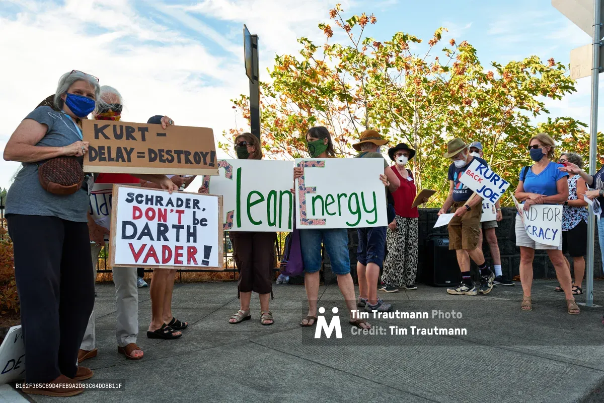 A diverse group of masked protesters gathers on a concrete sidewalk in Oregon City, holding handmade signs advocating for clean energy policies and criticizing Representative Kurt Schrader. The demonstrators, ranging from middle-aged to elderly participants, stand beneath autumn foliage under a partly cloudy sky, their colorful placards displaying messages like "Clean Energy" and "SCHRADER, DON'T be DARTH VADER!" The scene captures grassroots political activism in the Pacific Northwest, with protesters positioned along McLoughlin Promenade near the congressman's local office.