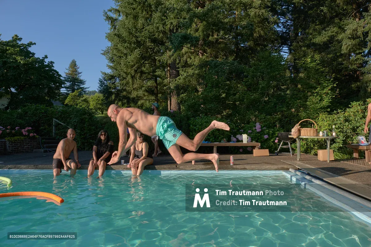 A shirtless man in mint green swim shorts launches into a backyard pool mid-leap, his body captured in dynamic motion against the serene backdrop of Newberg's Orchard Vibration. Three friends sit poolside watching the dive unfold, while towering evergreens and blooming hydrangeas frame this intimate summer gathering. The crystalline water below reflects dappled sunlight, creating an inviting aquamarine canvas for this moment of carefree abandon.