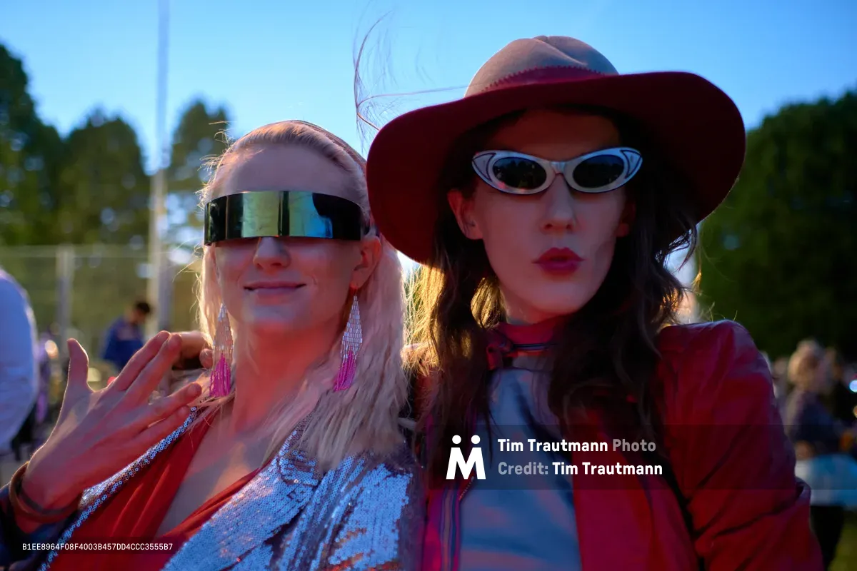 Two women pose in striking futuristic eyewear at Portland's Drop Out Prom Ride, part of the city's legendary Pedalpalooza cycling celebration in June 2019. The blonde woman sports sleek metallic wraparound sunglasses while her brunette companion wears vintage-style oval frames beneath a wide-brimmed hat, their faces catching the golden hour light. The outdoor gathering unfolds behind them in Colonel Summers Community Garden, where participants embrace whimsical costumes and alternative fashion for this beloved counter-cultural bike ride event.