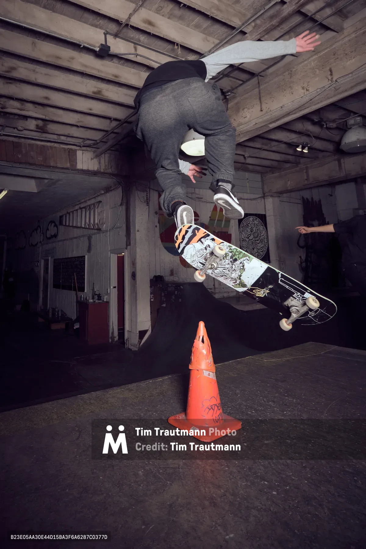 A skateboarder defies gravity in mid-air, soaring over a bright orange traffic cone inside the weathered confines of Commonwealth Skateboarding in Portland's Buckman neighborhood. The exposed wooden ceiling beams and graffitied walls create a gritty urban backdrop as the athlete executes a precise aerial maneuver, skateboard clutched firmly beneath gray-clad legs. Industrial lighting cuts through the basement-like atmosphere of this Southeast Portland skate haven, capturing the raw energy and creative spirit that defines the local skateboarding scene.