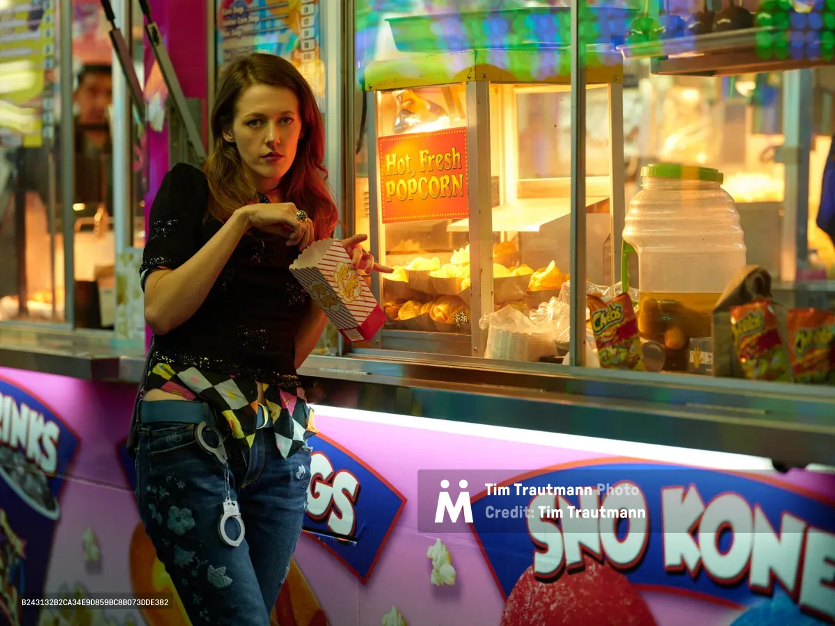 A young woman with auburn hair reaches into a glowing popcorn cart at the Clark County Fair in Vancouver, Washington. The warm golden light from the concession stand contrasts with the cool purple and blue neon of carnival rides spinning in the background. Her striped shirt and distressed jeans capture the casual energy of fairgoers, while the scene pulses with the electric atmosphere of late-night carnival revelry.