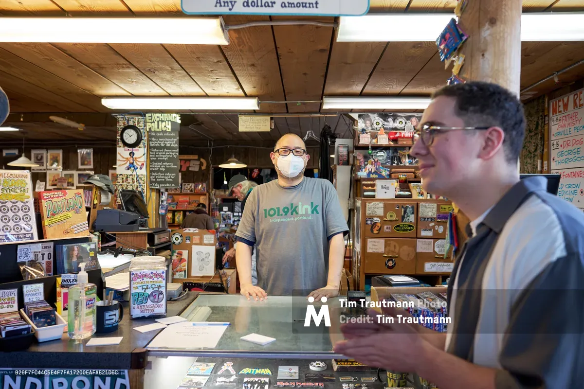 Inside Music Millennium on Portland's East Burnside during Record Store Day 2022, a masked store employee in a gray t-shirt stands behind the glass counter while engaging with a customer. The intimate record store atmosphere is illuminated by warm overhead lighting, revealing walls densely packed with vinyl records, vintage posters, and music memorabilia. The wooden beam ceiling and cluttered shelving create an authentic, lived-in feeling that captures the essence of Portland's independent music culture during this annual celebration.