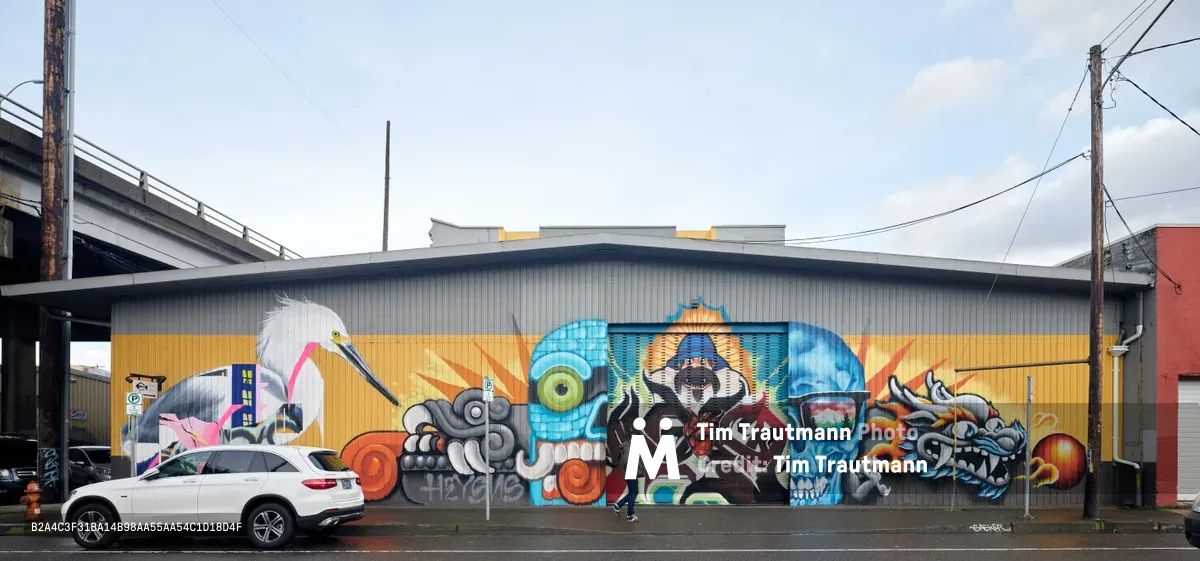 A sprawling street art mural covers the corrugated metal facade of an industrial warehouse in Portland's Central Eastside, featuring a dynamic collage of mythical creatures, skulls, and abstract forms in brilliant oranges, blues, and grays. The commissioned artwork by the Portland Street Art Alliance transforms the urban landscape beneath overcast Pacific Northwest skies, while a white SUV passes along the quiet street lined with power lines and industrial infrastructure. The mural's fantastical imagery—including a prominent white crane, mechanical elements, and surreal characters—creates a striking contrast against the building's utilitarian architecture.