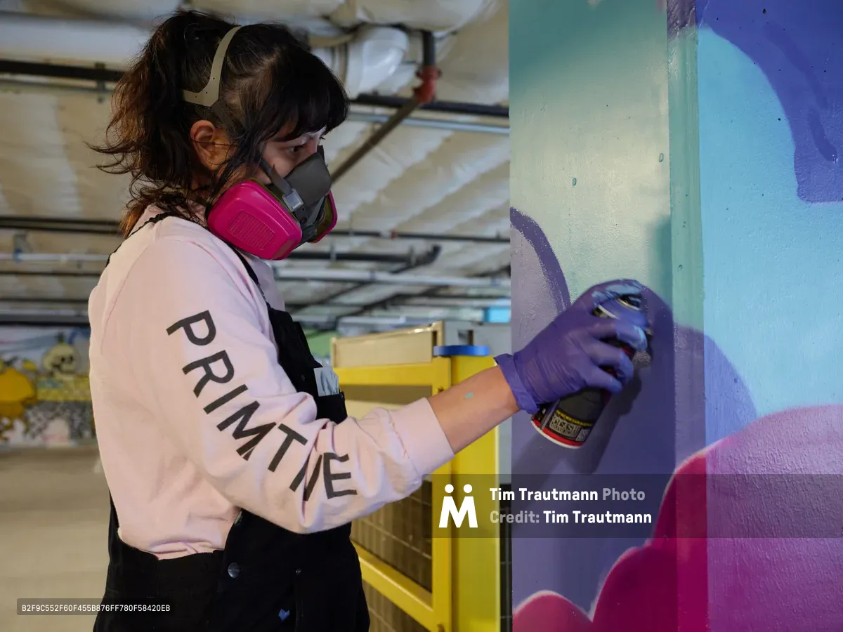 A focused muralist wearing protective gear and purple gloves carefully applies spray paint to a vibrant wall inside Clay Creative's parking garage in Southeast Portland. The artist, dressed in a pink 'PRIMETIME' sweatshirt and respirator mask, works methodically on flowing abstract forms in blues and purples. Industrial ceiling fixtures cast even light across the underground creative space, while yellow structural elements provide stark contrast to the emerging artwork.