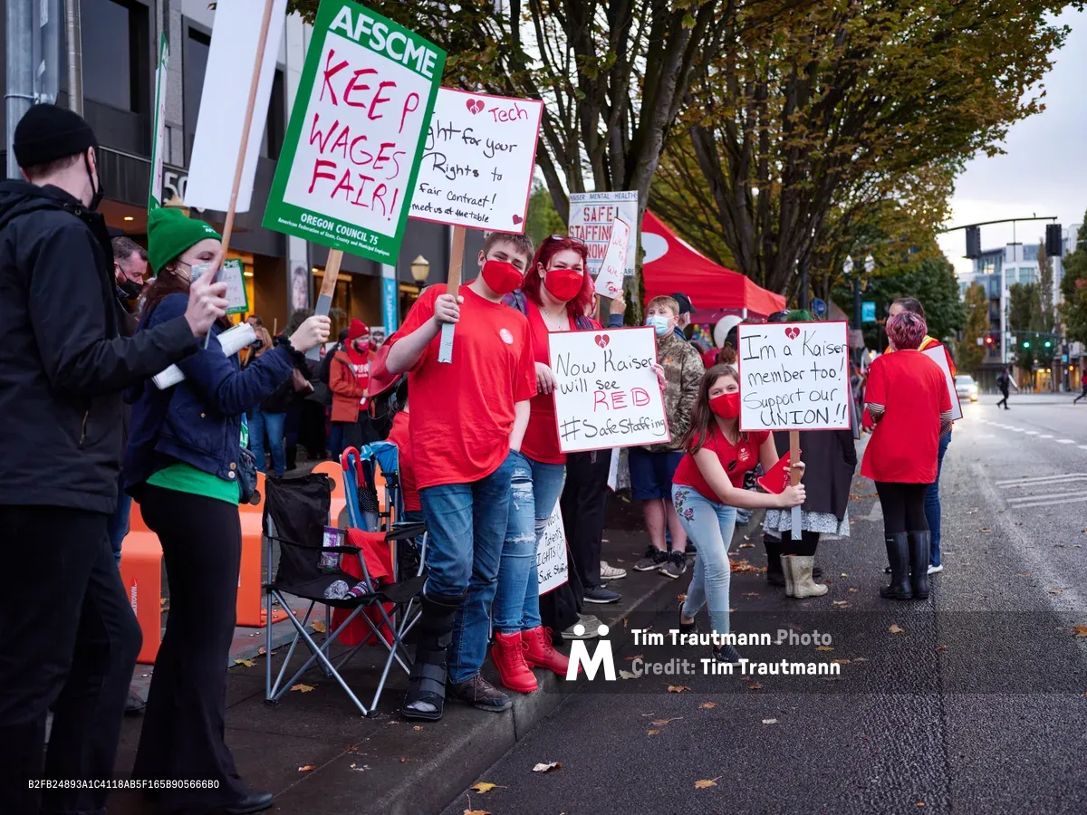 Healthcare workers in crimson masks and shirts gather on Northeast Multnomah Street outside Kaiser Permanente Tower, their handmade signs demanding fair wages and union recognition piercing the overcast Portland evening. The AFSCME banners and personal placards create a sea of red against the urban backdrop, while folding chairs and determined faces signal a prolonged demonstration. Autumn leaves scatter across wet pavement as protesters maintain their vigil beneath the towering medical complex, their collective voice rising in the Lloyd District's fading light.