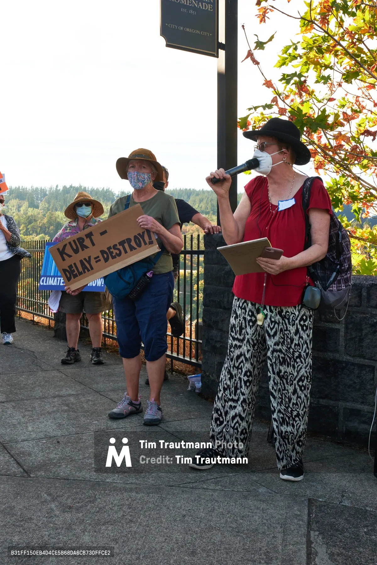 Masked protesters gather beneath autumn foliage at McLoughlin Promenade in Oregon City, where a woman in red speaks into a microphone while others hold handmade signs demanding climate action. The scene unfolds in golden afternoon light, with demonstrators positioned near Representative Kurt Schrader's office, their urgent message carried against the backdrop of forested hills and seasonal maple leaves in warm transition colors.