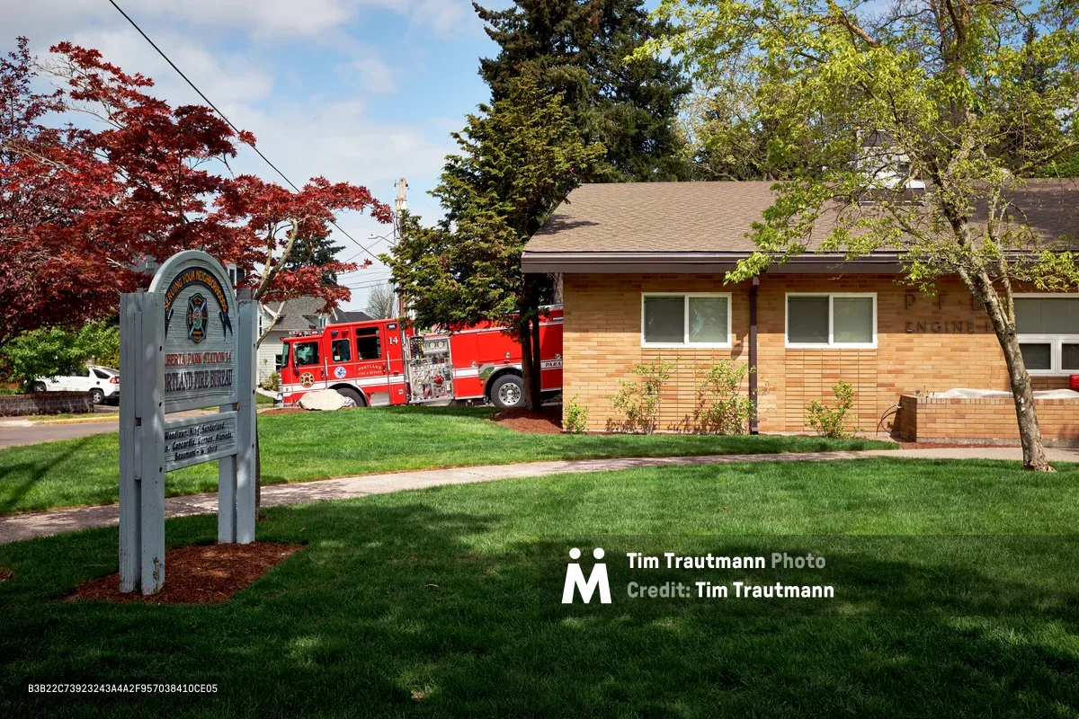 A bright red fire engine sits stationed outside Portland Fire & Rescue Station 14 on Northeast Killingsworth Street in the Alberta neighborhood. The modern brick fire station is framed by vibrant spring foliage, including deep crimson Japanese maples and fresh green deciduous trees. A professional white sign stands prominently in the manicured lawn, marking this community emergency services facility against a backdrop of scattered clouds and residential homes.