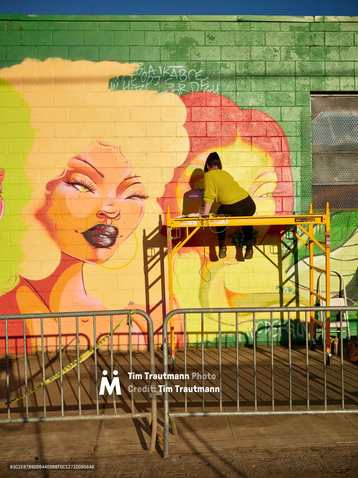 Multidisciplinary artist Vilatose works from a yellow scaffolding platform, adding details to Jardín Cubana, a vibrant mural featuring three female figures on the brick wall at Donny Vegas in Portland's Alberta Arts District. The mural bursts with warm yellows, coral pinks, and emerald greens against the industrial backdrop of metal barriers and urban streetscape. Late afternoon light bathes the scene as the artist, dressed in a mustard yellow sweatshirt, carefully refines the figurative composition with concentrated precision.