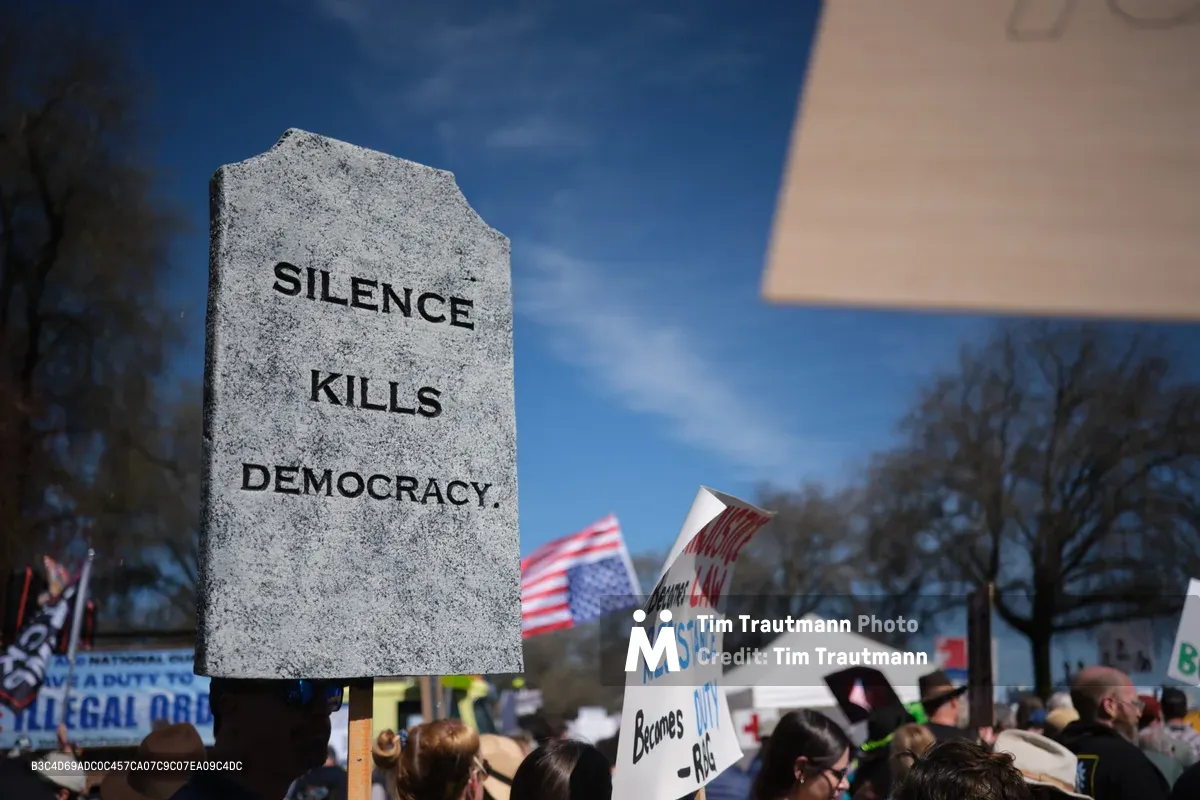 A weathered granite tombstone bearing the stark inscription 'SILENCE KILLS DEMOCRACY' towers above a sea of protesters gathered in Portland's Chinatown district. The mock gravestone, held aloft against a crisp blue sky dotted with white clouds, serves as the focal point of the March 2026 No Kings demonstration. American flags flutter among the crowd while various protest signs create a mosaic of dissent, including visible text about resistance and legal obligations.