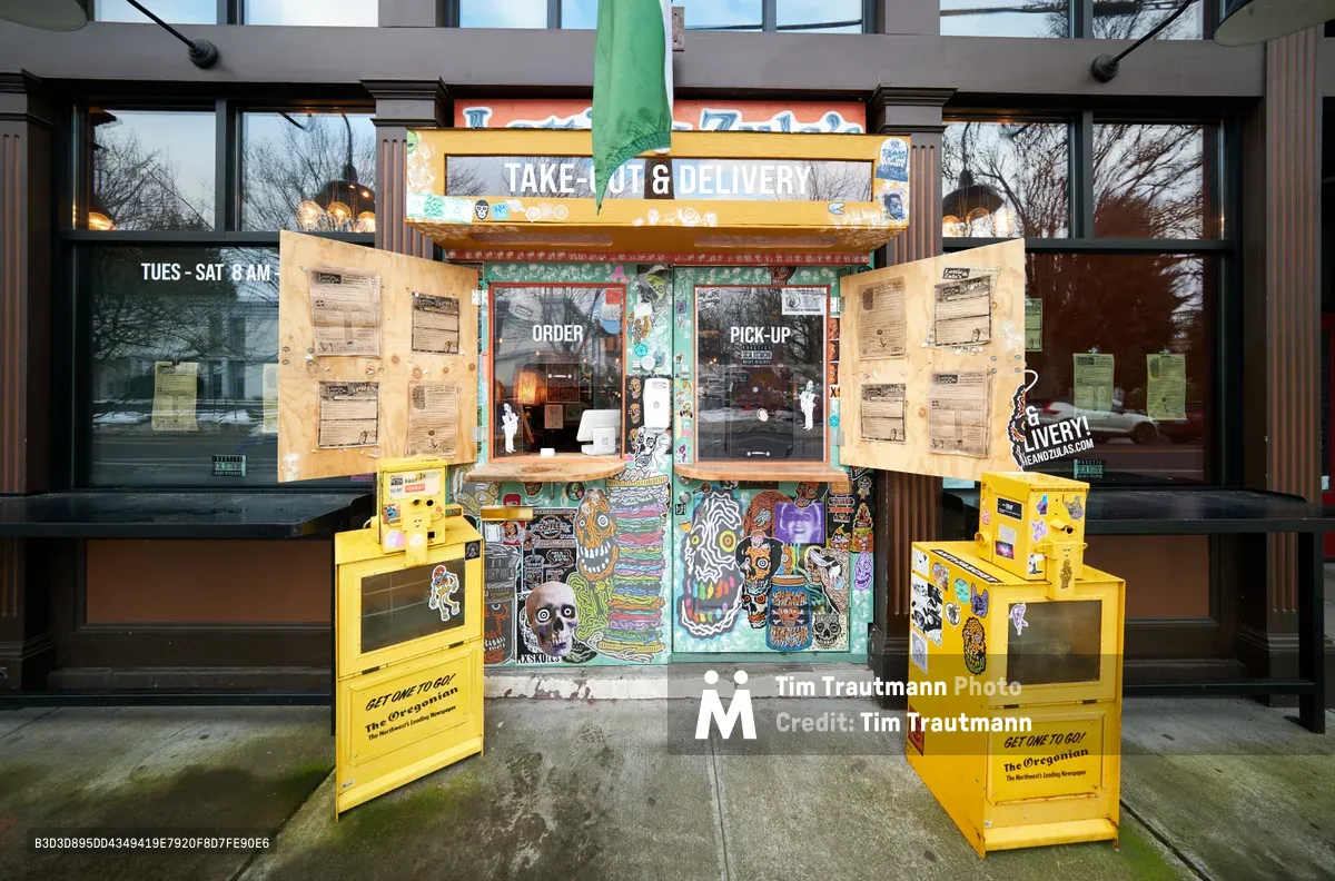 A custom-built takeout station integrates seamlessly into the main entrance of Lottie and Zula's restaurant in Portland's Eliot neighborhood, flanked by bright yellow Oregonian newspaper boxes. The purposefully constructed wooden counter, adorned with vibrant street art featuring skulls and psychedelic patterns, bears witness to the restaurant industry's adaptive spirit during COVID-19. Behind rain-streaked windows, the darkened interior hints at a business transformed, while menu boards and pickup instructions create an improvised commerce hub on the sidewalk.
