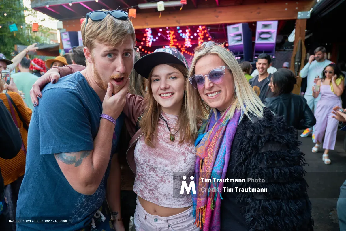 Three festival-goers embrace in a moment of pure joy at White Owl Social Club's Your Sunday Best day party in Portland, Oregon. The trio - a bearded man in blue flannel making a playful gesture, a young woman in bohemian attire with a black cap, and a blonde woman draped in a vibrant shawl and black feathers - radiate infectious energy against the backdrop of string lights and warm wooden architecture. The outdoor venue pulses with summer festival atmosphere as other revelers dance and socialize in the golden afternoon light.