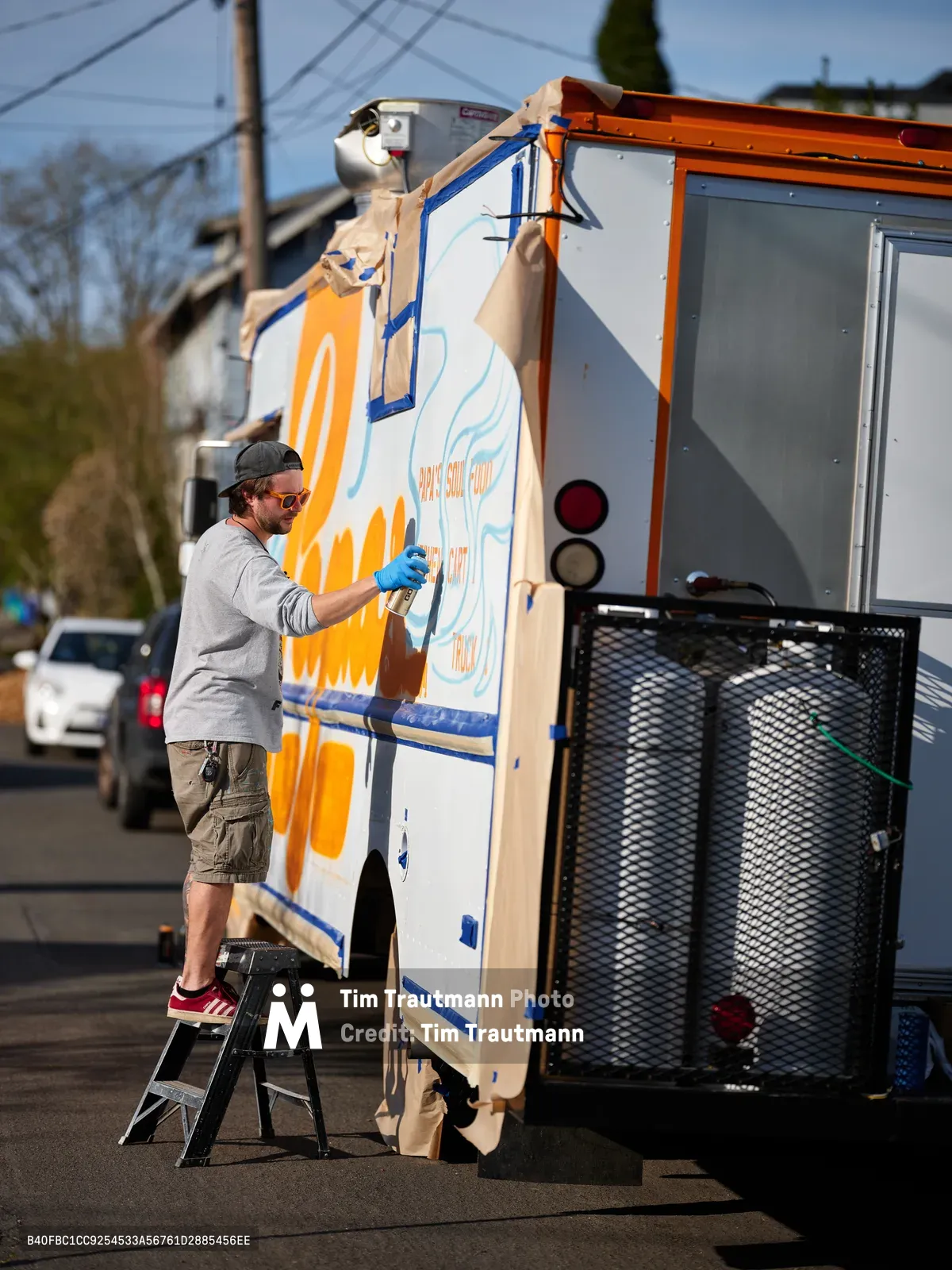 A bearded muralist in casual attire stands on a stepladder, spray-painting vibrant orange and blue designs onto the side of a white food truck parked along Northeast 9th Avenue in Portland's King neighborhood. The afternoon light illuminates his concentrated work as protective plastic sheeting flutters around the vehicle's windows. Power lines and residential buildings create an urban backdrop typical of Portland's eclectic food truck culture.