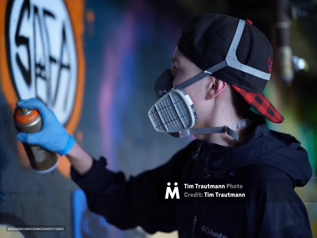 A young artist wearing a respirator mask and baseball cap carefully applies spray paint to a vibrant mural in the industrial confines of Clay Creative's parking garage in Portland's Hosford-Abernethy neighborhood. The atmospheric lighting catches swirls of aerosol mist as colorful graffiti blooms across concrete walls, transforming the utilitarian space into an underground canvas. Blue-gloved hands guide the spray can with practiced precision while neon hues pierce through the shadowy garage interior.