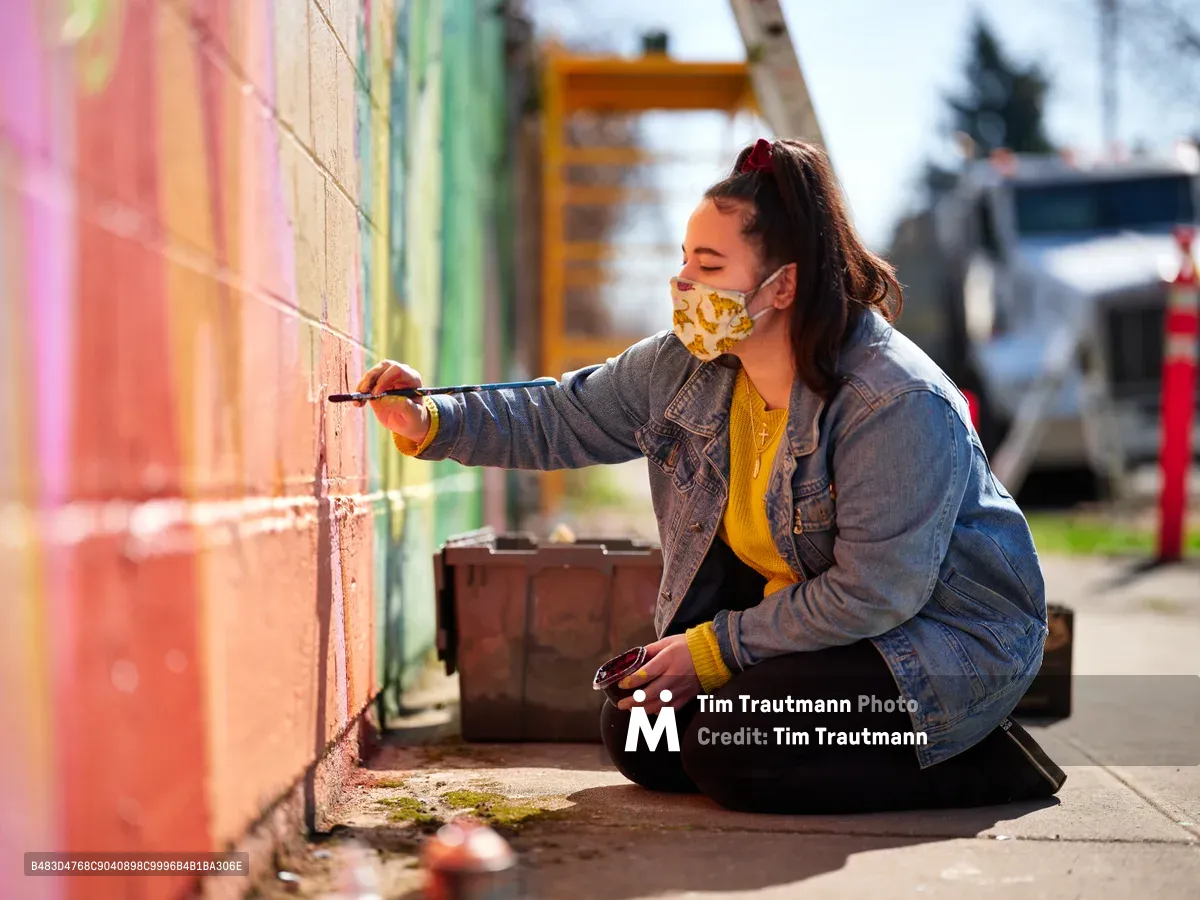 Multidisciplinary artist Villatose sits cross-legged on the sidewalk, brush in hand, adding intricate details to her 'Jardín Cubana' mural featuring three female figures along Alberta Street in Portland's arts district. Golden afternoon light bathes the scene as she works methodically on the colorful wall, her floral face mask and mustard sweater creating a striking contrast against the weathered denim jacket. The composition captures the intimate concentration of street art creation, with urban elements softly blurred in the background.
