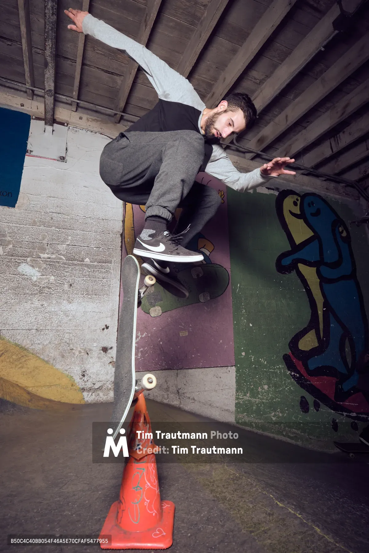 A bearded skateboarder in gray sweatpants and white hoodie executes a trick over a red traffic cone in the gritty underground confines of Commonwealth Skateboarding in Portland's Buckman neighborhood. The raw concrete walls are adorned with colorful graffiti art, while weathered wooden ceiling beams cast dramatic shadows across the subterranean skate sanctuary. His focused expression and extended arms capture the precise balance required as he navigates the urban obstacle course beneath the city streets.