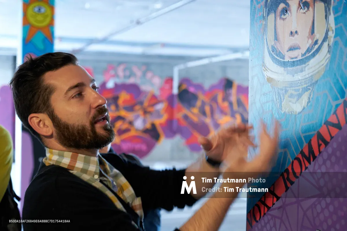 Tomás Valladares of the Portland Street Art Alliance gestures expressively while explaining the intricate details of a Voxx Romana mural in Southeast Portland's Hosford-Abernethy neighborhood. His animated discussion unfolds against a vibrant backdrop of colorful street art, where the astronaut-themed mural's blues and whites contrast with the purple and orange graffiti beyond. The intimate educational moment captures the passion of urban art advocacy in Clay Creative's artistic environment.