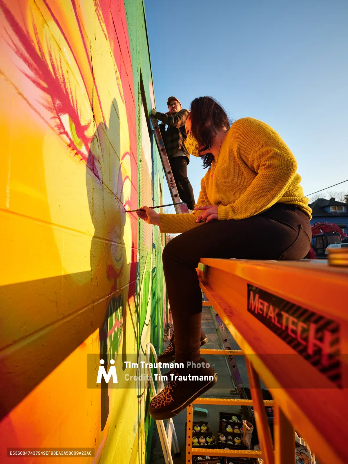 Multidisciplinary artist Vilatose works on the dynamic mural Jardín Cubana from an orange Metaltec scissor lift on Alberta Street in Portland's arts district. The late afternoon light bathes the scene in warm golden tones as the artist, wearing a mustard yellow sweater, carefully applies paint to the bold composition depicting three female figures spanning bright yellows, coral pinks, and lime greens. A second collaborator assists from above while the Alberta Arts District's creative energy pulses through this moment of urban art creation.