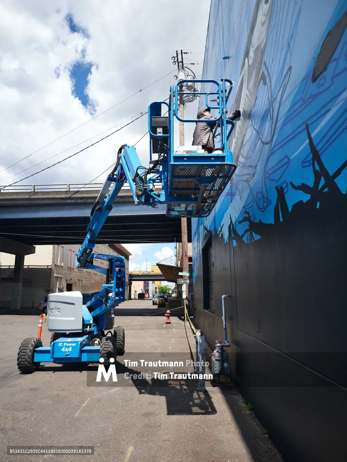 Jeremy Nichols and Devin Finley work from a blue hydraulic lift to paint a vibrant blue mural on an industrial building wall in Southeast Portland. The scene captures the intersection of urban creativity and industrial infrastructure, with the artists elevated against a backdrop of power lines, concrete structures, and the characteristic overcast Pacific Northwest sky. Safety cones mark the work zone while the lift's mechanical precision contrasts with the flowing organic forms taking shape on the wall.