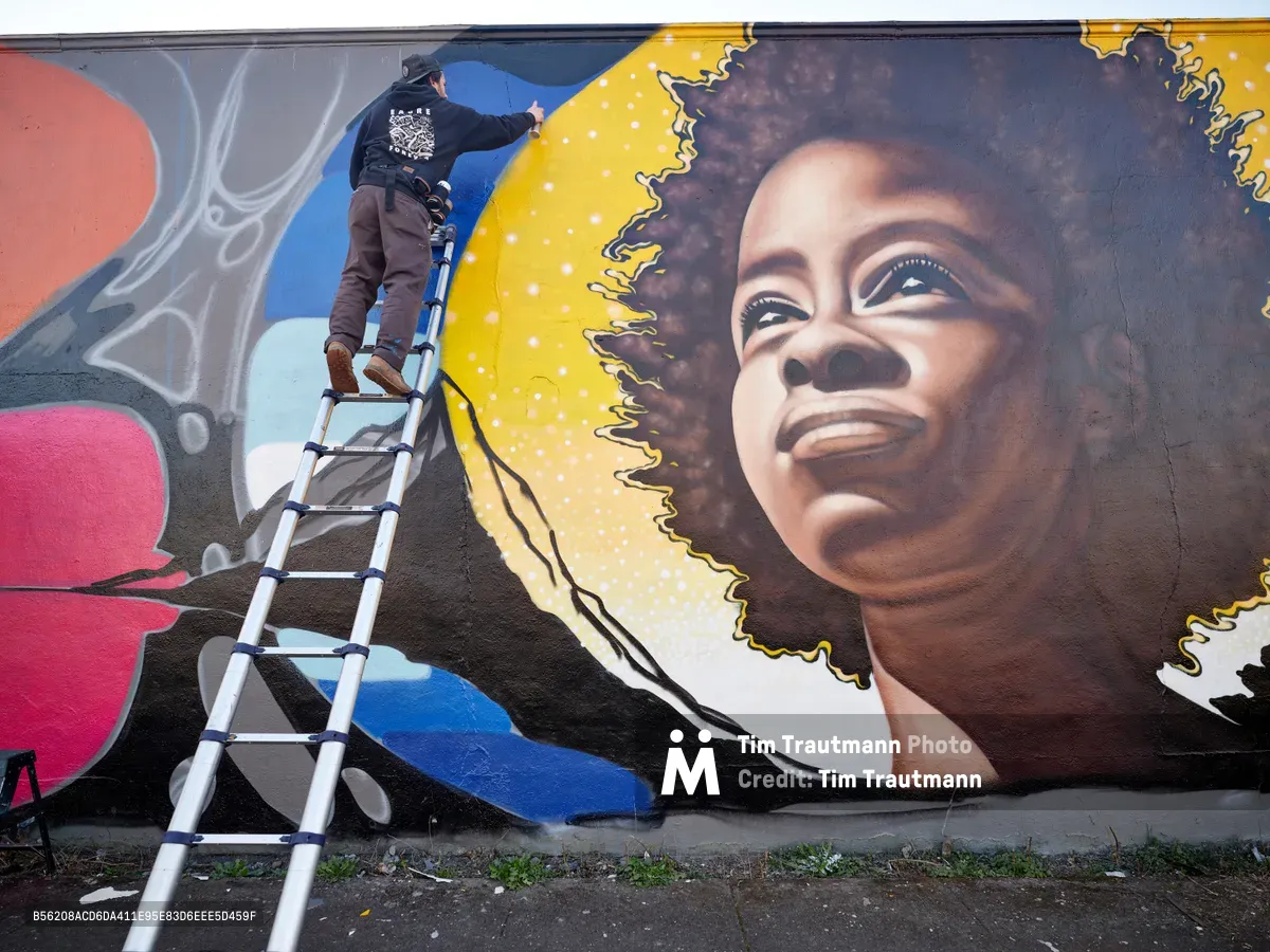 Street artist Travis Fields, known as Campographic, meticulously works from an aluminum ladder to paint a vibrant portrait of poet Amanda Gorman on a concrete wall in Portland's Alberta district. The mural captures Gorman's likeness with remarkable detail, featuring her natural hair crowned with a golden halo against a backdrop of swirling colors and abstract elements. The scene unfolds on Northeast Killingsworth Street, where the artist's concentrated effort transforms the urban canvas into a celebration of literary achievement.