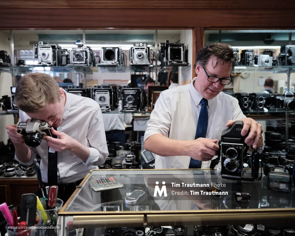 In the intimate confines of Blue Moon Camera & Machine, Portland's legendary camera haven, Mike Knight and a colleague work with reverent concentration behind a gleaming glass counter. The warm tungsten light illuminates their careful examination of vintage equipment while towering displays of classic cameras—from twin-lens reflex models to large format view cameras—create a museum-like backdrop. The scene captures the quiet dedication of analog photography's guardians, their focused gestures speaking to decades of expertise amid this treasure trove of mechanical precision.