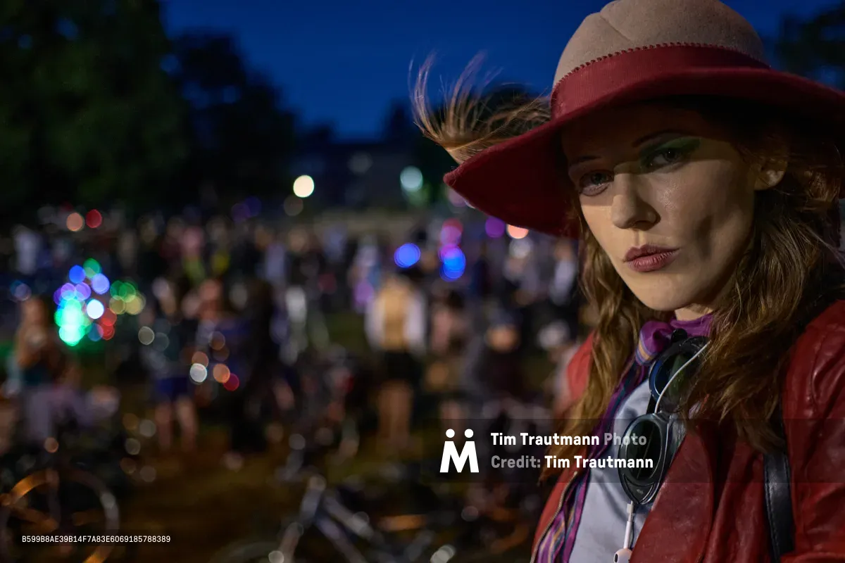 A young woman in a wide-brimmed burgundy hat gazes thoughtfully into the camera during Portland's unconventional Drop Out Prom Ride event at Colonel Summers Community Garden. The twilight hour bathes her face in soft, dramatic light while bokeh circles from distant festival lights create a dreamy backdrop against the deep blue evening sky. Her vintage-inspired outfit and contemplative expression capture the whimsical rebellion of this alternative prom celebration, part of Portland's annual Pedalpalooza cycling festival.