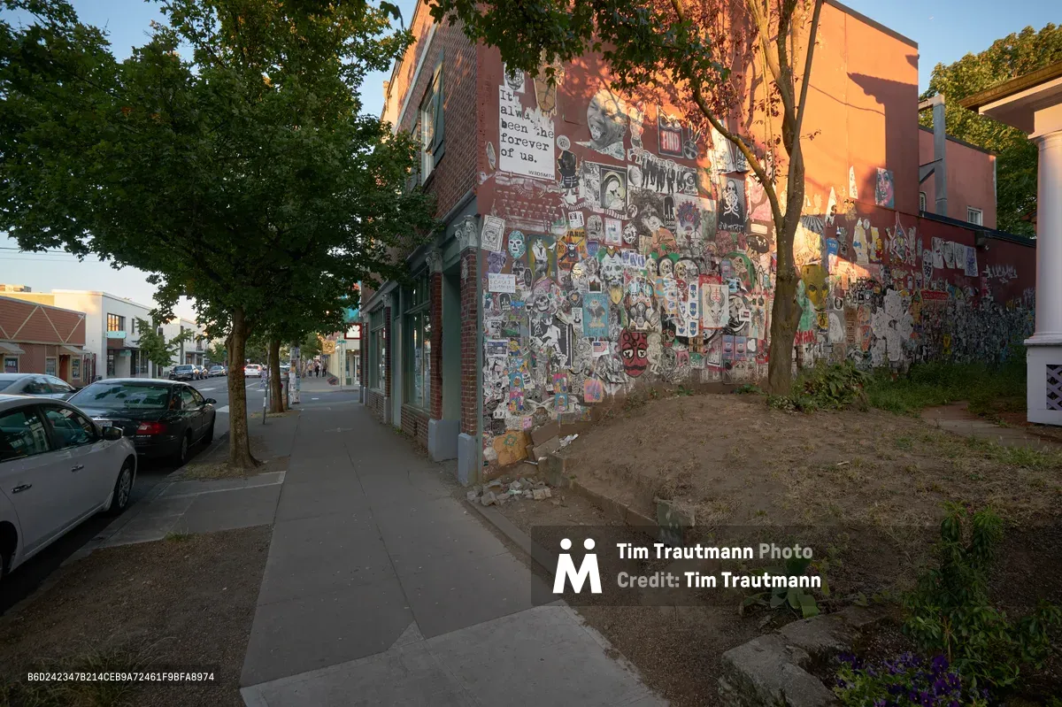 Golden hour light bathes a weathered brick corner building adorned with hundreds of colorful stickers, posters, and street art along Northeast Alberta Street in Portland's Alberta Arts District. The eclectic collage creates a vibrant tapestry of pop culture references, local art, and urban expression against the warm red brick facade. Leafy trees frame the scene while parked cars and sidewalk hint at the neighborhood's lived-in character.
