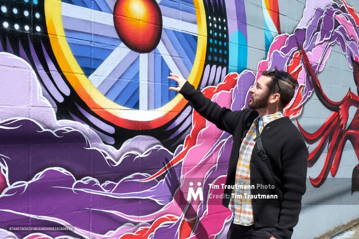 Tomás Valladares of the Portland Street Art Alliance reaches toward a vibrant mural during an interpretive tour in Portland's Central Eastside district. The dramatic wall art features swirling purple and pink forms surrounding a luminous orange orb, creating a dynamic cosmic landscape against the concrete surface. Valladares, dressed in a dark sweater and plaid shirt with a messenger bag, gestures enthusiastically as he shares the story behind the artwork. The scene captures the intersection of community education and urban artistry on Southeast 11th Avenue.