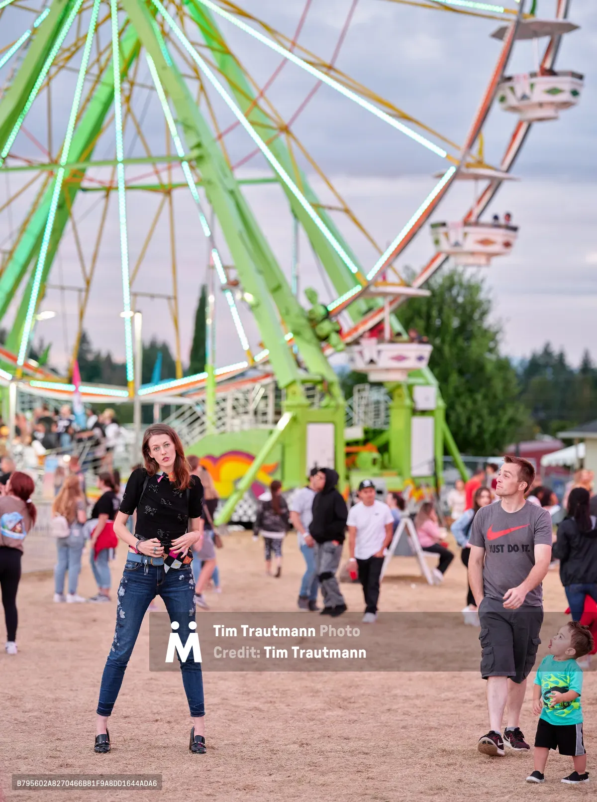 Under the soft glow of dusk at Vancouver's Clark County Fair, a young woman in a black patterned top and distressed jeans strikes a confident pose in the foreground, while families with children enjoy the carnival atmosphere behind her. The towering lime-green Ferris wheel dominates the frame, its passenger gondolas catching the warm evening light as fairgoers mill about on the dusty midway grounds. The scene captures the nostalgic charm of Pacific Northwest summer traditions, where generations gather for rides, games, and community celebration.
