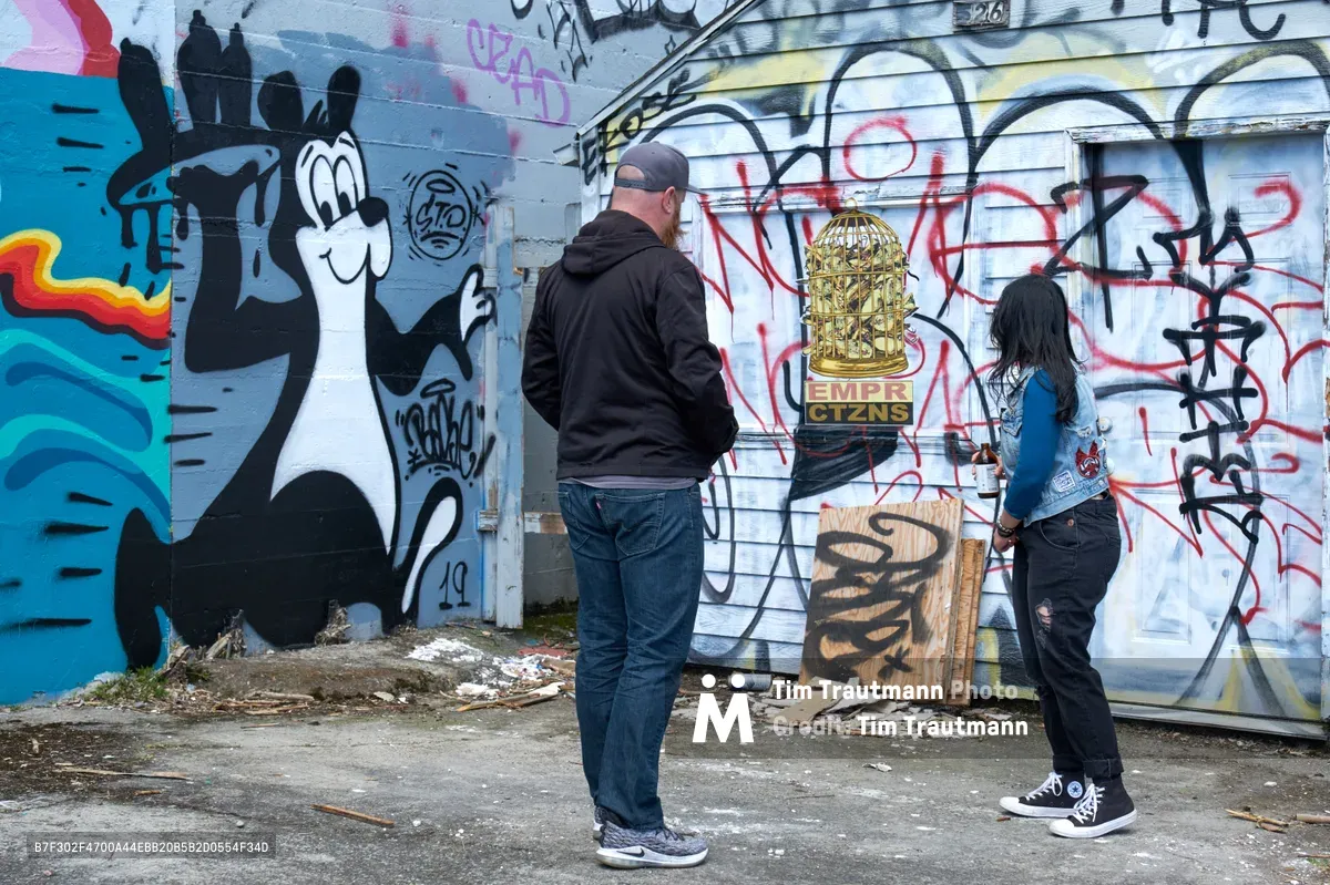Two visitors stand before a vibrant mural wall in Portland's Central Eastside during a guided street art tour. The weathered concrete surface displays layers of graffiti including a whimsical cartoon character, ornate lettering, and colorful geometric forms. The tour guide and participant examine the urban artwork amid scattered debris on the industrial pavement, capturing the raw authenticity of this evolving neighborhood's creative expression.