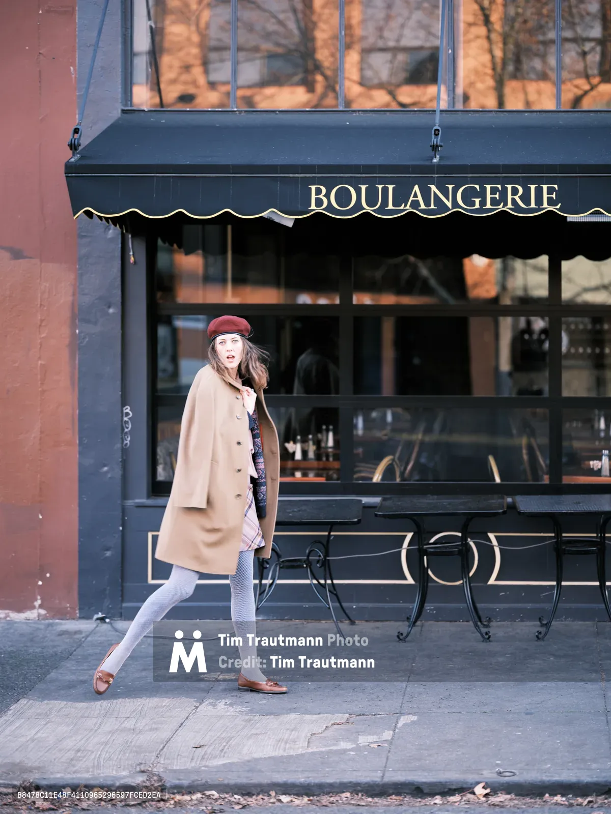 A young woman in a burgundy beret and camel wool coat strides past a charming French bakery on Northeast Alberta Street in Portland's Alberta district. The golden morning light catches the storefront's navy awning emblazoned with 'BOULANGERIE' in elegant serif lettering, while bare winter branches create delicate shadows across the scene. Her mid-stride pose and surprised expression suggest she's been caught in a candid moment of urban exploration, embodying the effortless style of Portland's creative neighborhoods.