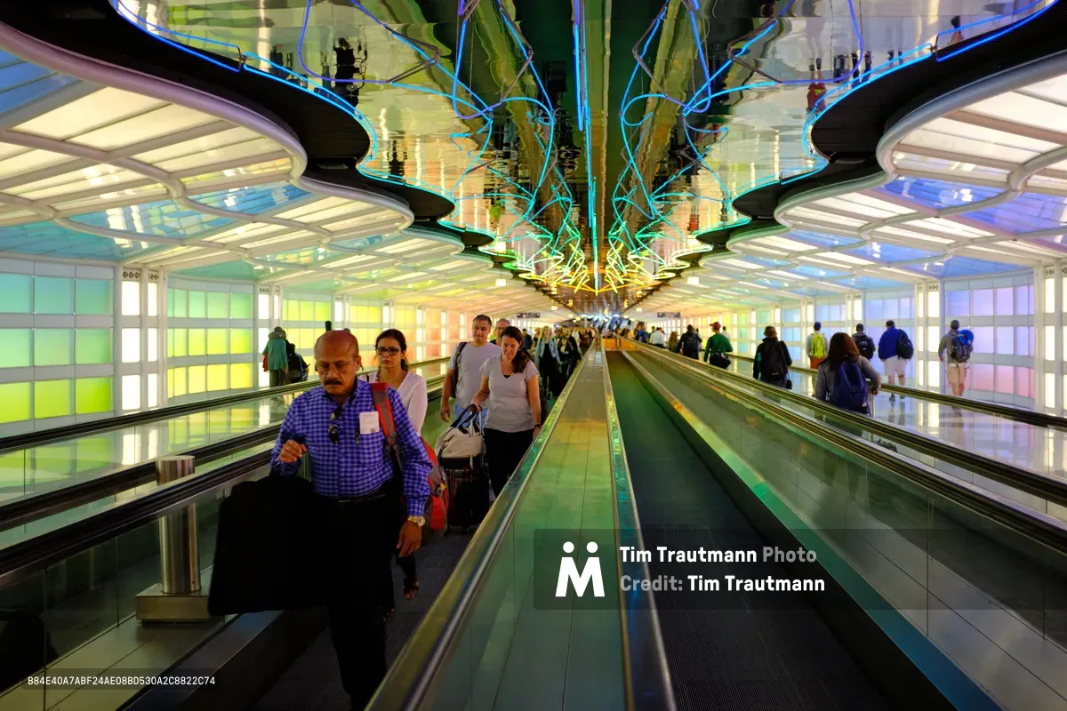 Travelers glide through the ethereal rainbow tunnel of Chicago O'Hare International Airport's pedestrian walkway, where curved glass walls pulse with vibrant neon hues from green to purple. An elderly man in a blue checkered shirt leads the procession down the moving walkway, while silhouettes of fellow passengers create a sense of purposeful movement against the kaleidoscopic backdrop. The futuristic architecture transforms the mundane act of airport transit into a journey through a luminous portal, embodying the limitless possibilities that air travel represents.
