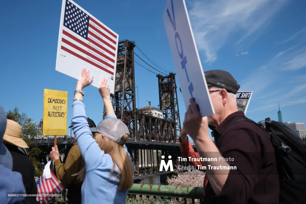 Protesters gather beneath Portland's Steel Bridge on a brilliant spring day, raising American flags and yellow signs declaring "Democracy Not Autocracy" against the industrial lattework of the historic lift bridge. The scene captures citizens exercising their First Amendment rights with the Willamette River waterfront as backdrop, while the bridge's towering steel framework creates dramatic geometric patterns against the azure sky. Demonstrators in casual attire hold their signs high, their message echoing across the water toward downtown Portland's skyline visible in the distance.