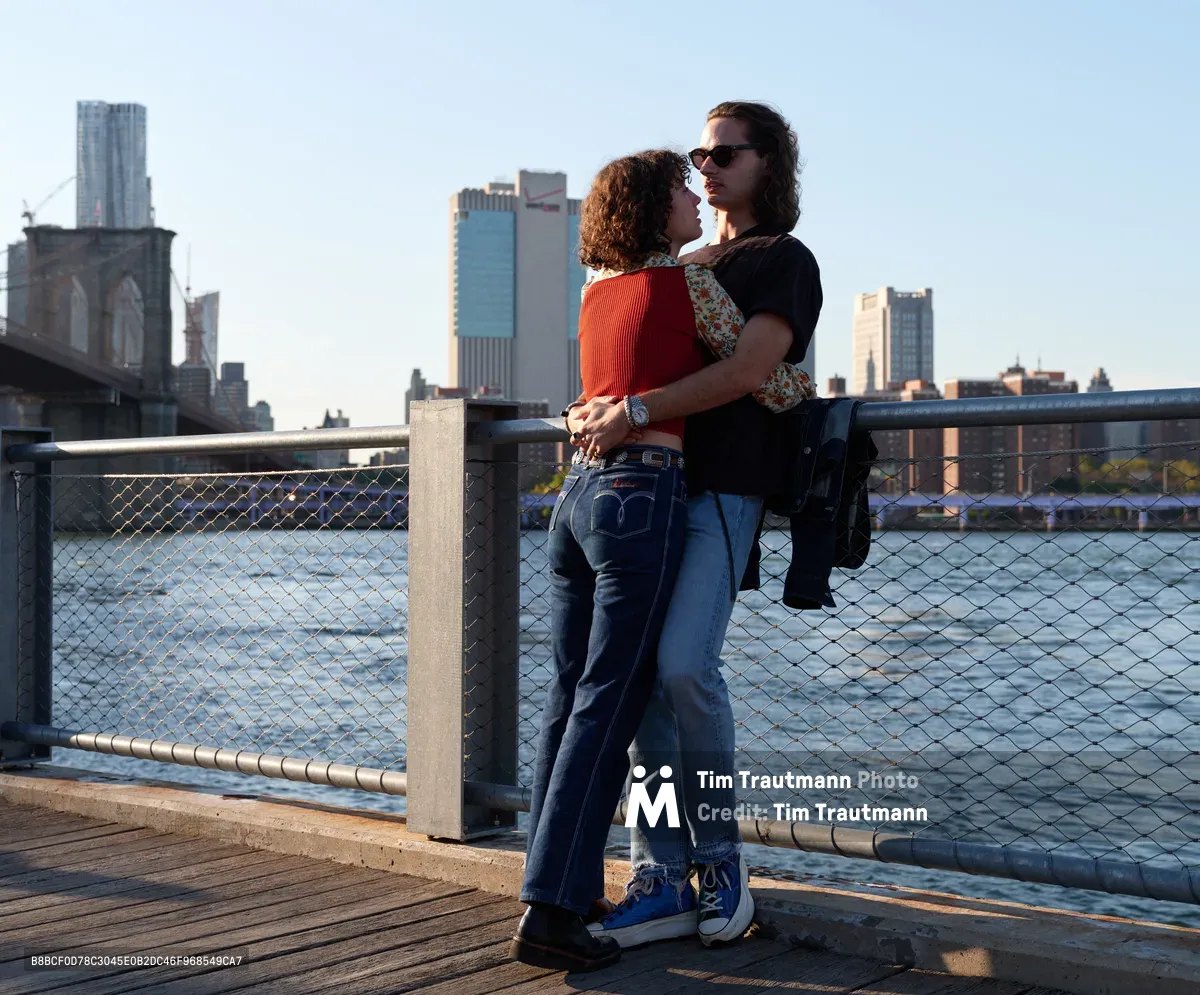 A couple shares an intimate embrace along the East River waterfront in DUMBO, Brooklyn, with the iconic Manhattan skyline creating a dramatic backdrop. The woman in a vibrant orange sweater and the man in dark clothing stand close against the chain-link railing as golden hour light bathes the scene. The Brooklyn Bridge's Gothic arches frame the left side while modern skyscrapers pierce the soft blue sky, creating a quintessential New York romance scene.