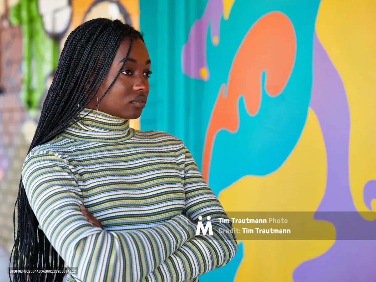 Naomi Likayi stands confidently with crossed arms before her vibrant street mural in Portland's Alberta Arts District. Her long box braids cascade over a striped mock-neck sweater as she gazes thoughtfully over her shoulder. The explosive backdrop features bold organic shapes in turquoise, coral orange, sunny yellow, and purple, creating a dynamic contrast against the urban setting. Soft natural light illuminates her face, capturing a moment of artistic accomplishment and quiet pride.