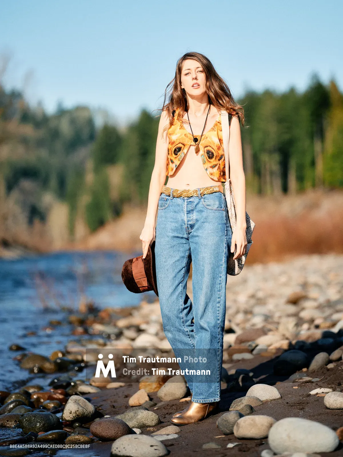 A young woman stands contemplatively among smooth river stones along the Floodplain Trail in Multnomah County, Oregon. She wears vintage-inspired denim and a whimsical owl-print top, her flowing hair catching the golden hour light. The serene riverbed setting creates an intimate portrait of connection with nature, while her leather bag and bohemian accessories suggest a wanderer's spirit. Gentle waters lap at the rocky shore behind her, framed by Oregon's lush forest landscape.