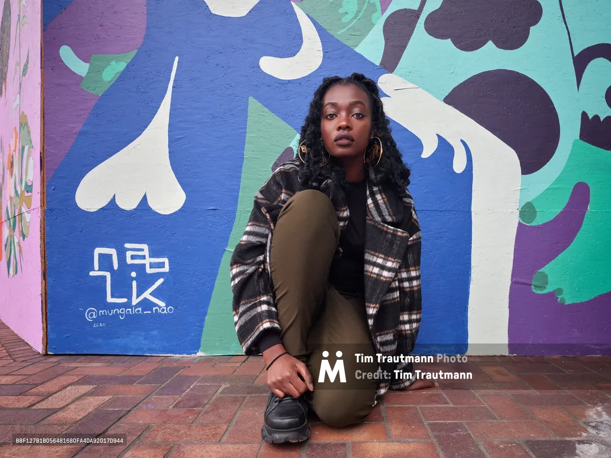 Artist Naomi Likayi sits contemplatively before her vibrant street mural at Portland's World Trade Center, her dark curls framing a direct, confident gaze. The abstract artwork behind her flows in bold blues, purples, and whites across the concrete wall, while warm afternoon light illuminates her olive jumpsuit and plaid flannel jacket. Her relaxed pose on the brick pavement suggests both ownership of her creative work and connection to the urban environment that serves as her canvas.