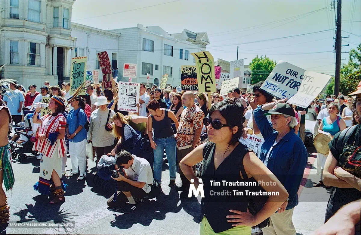 Sunlight bathes a diverse crowd of Mission District residents as they gather on Bryant Street, their handmade protest signs creating a forest of resistance against displacement. The warm afternoon light illuminates faces of determination—elderly activists in bucket hats stand alongside young mothers and working-class neighbors, their signs reading "Our City Not For Sale" and "Home People Over Profit." Victorian architecture looms behind the demonstrators, while power lines slice across an overcast sky, framing this snapshot of grassroots activism in San Francisco's historically Latino neighborhood.