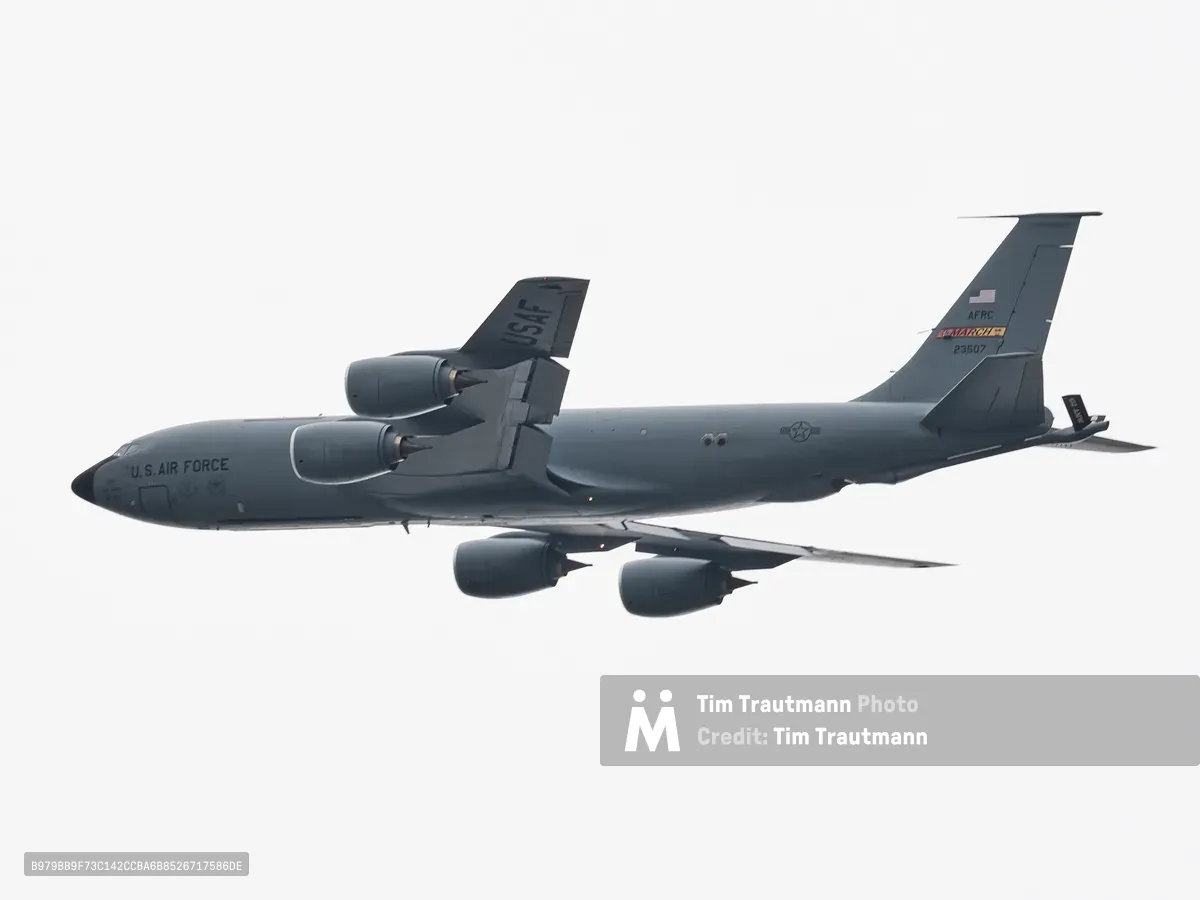 A U.S. Air Force Boeing KC-135 Stratotanker aerial refueling aircraft photographed in flight from below against a flat, overcast white sky. The aircraft is painted in overall grey and bears "U.S. AIR FORCE" lettering on the fuselage. "USAF" is stenciled on the vertical stabilizer, with "AFRC" and "MARCH" unit markings and the tail number 23507 visible near the tail. Four podded jet engines are mounted under the swept wings. The aircraft's boom refueling equipment is visible extending from the rear of the fuselage. A U.S. national insignia roundel appears on the fuselage. The tight telephoto framing isolates the aircraft cleanly against the grey-white sky.