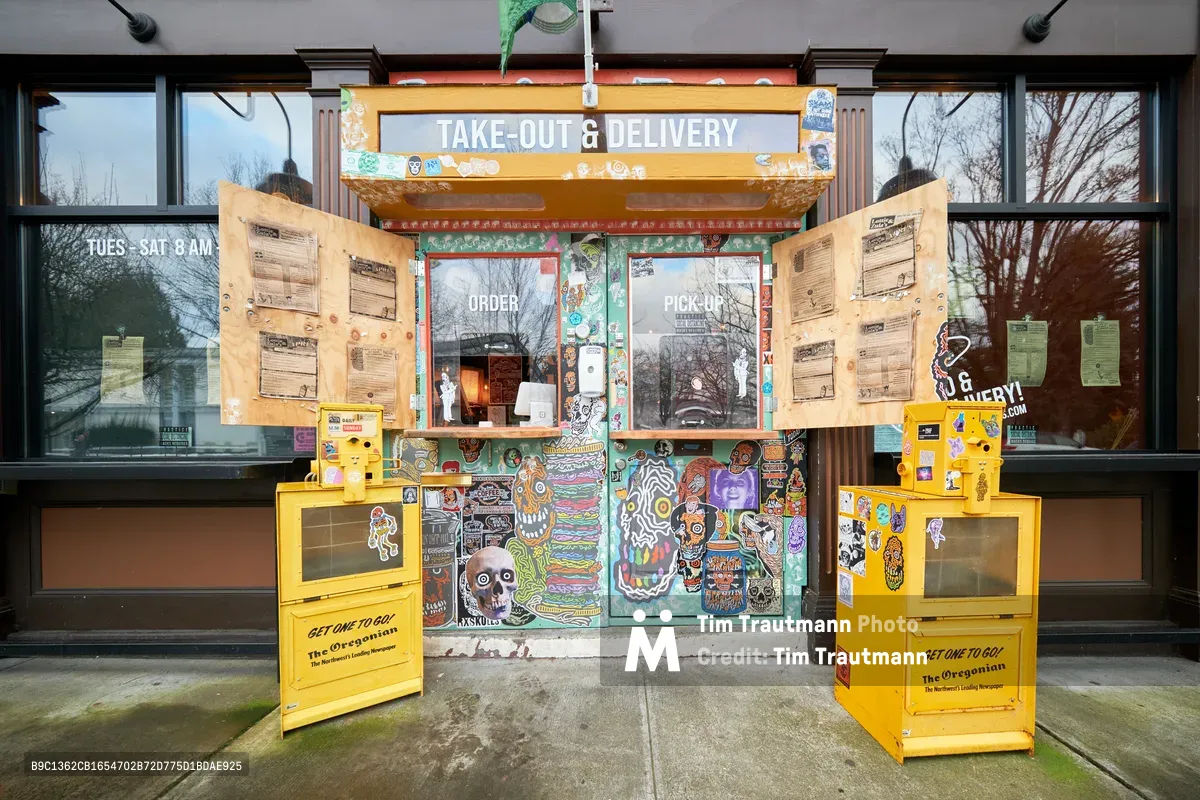 A custom-built takeout counter transforms the main entrance of Lottie and Zula's restaurant in Portland's Eliot neighborhood during the COVID-19 pandemic. Bright yellow newspaper stands flank a colorful graffiti-adorned storefront, where handwritten menus are taped to windows beside bold "ORDER" and "PICK-UP" signage. The purpose-built counter installation captures the resourceful spirit of local businesses pivoting to survive, with overcast winter light filtering through bare trees reflected in the glass facade.