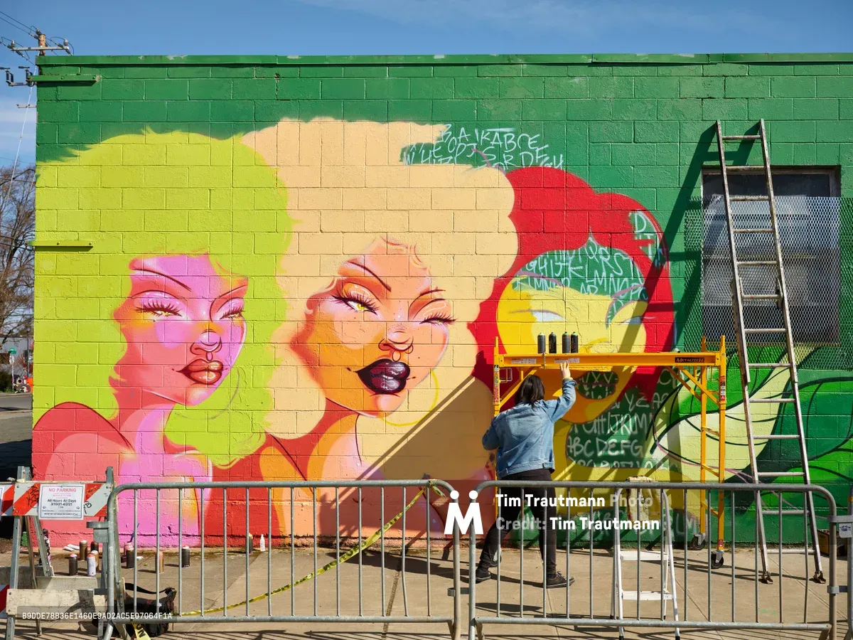 Multidisciplinary artist Vilatose applies vibrant pigments to 'Jardín Cubana,' a larger-than-life portrait featuring three female figures on Northeast Alberta Street, transforming a green cinderblock wall into a kaleidoscope of feminine faces rendered in electric pinks, corals, and golden yellows. Working from scaffolding beneath Portland's crystalline winter sky, the artist adds delicate details to expressive eyes and glossy lips while construction barriers frame the creative process. The emerging mural radiates pop art sensibilities against the industrial backdrop of the Alberta Arts District.