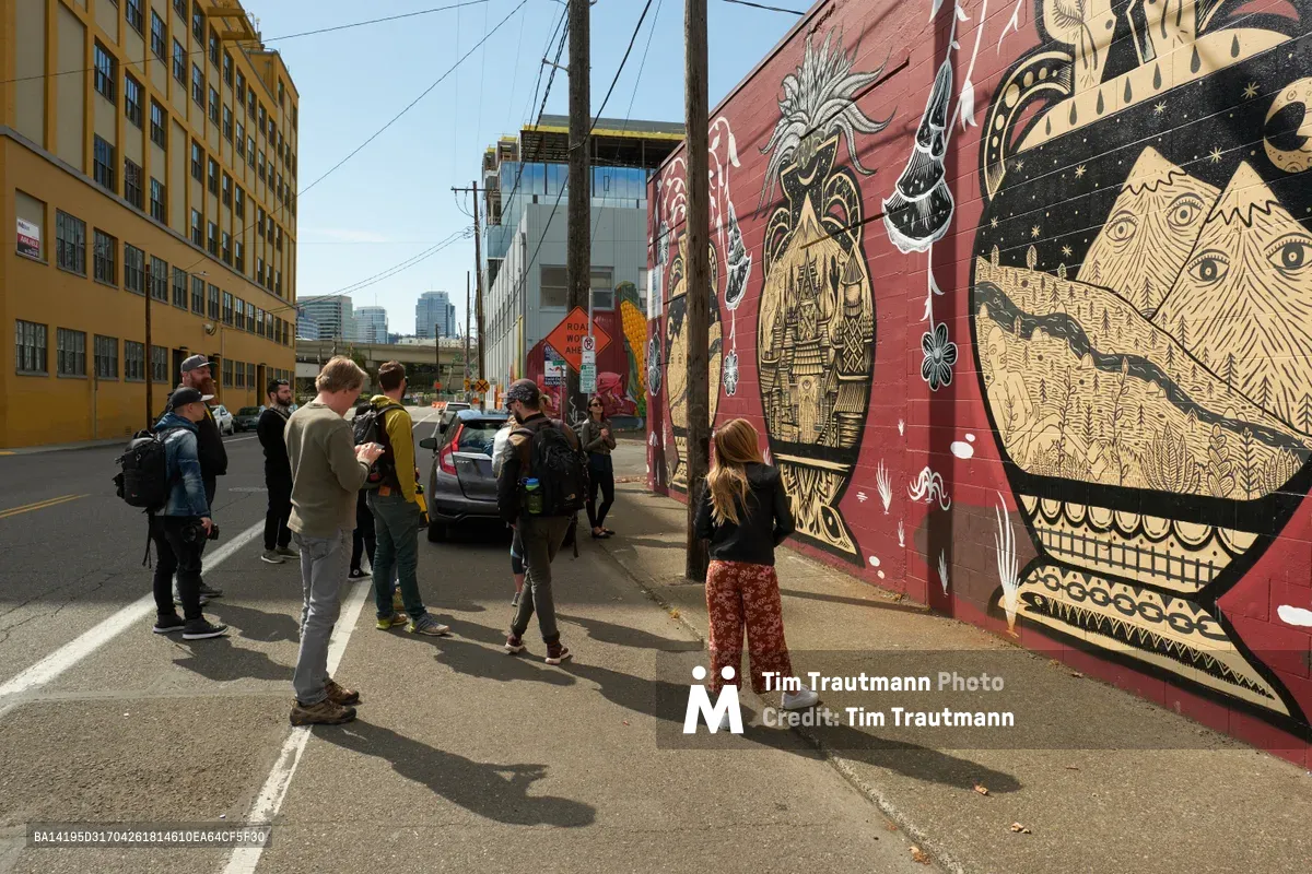 A diverse group of street art enthusiasts gathers on Southeast Stark Street in Portland's Central Eastside, their shadows stretching across weathered asphalt as they contemplate an intricate mural. The towering artwork features mystical creatures and celestial motifs in warm terracotta and cream tones, while the industrial streetscape of yellow brick buildings and power lines frames this moment of cultural discovery. The afternoon light creates dramatic silhouettes of the tour participants, each absorbed in studying the elaborate wall art that transforms this urban corridor into an open-air gallery.