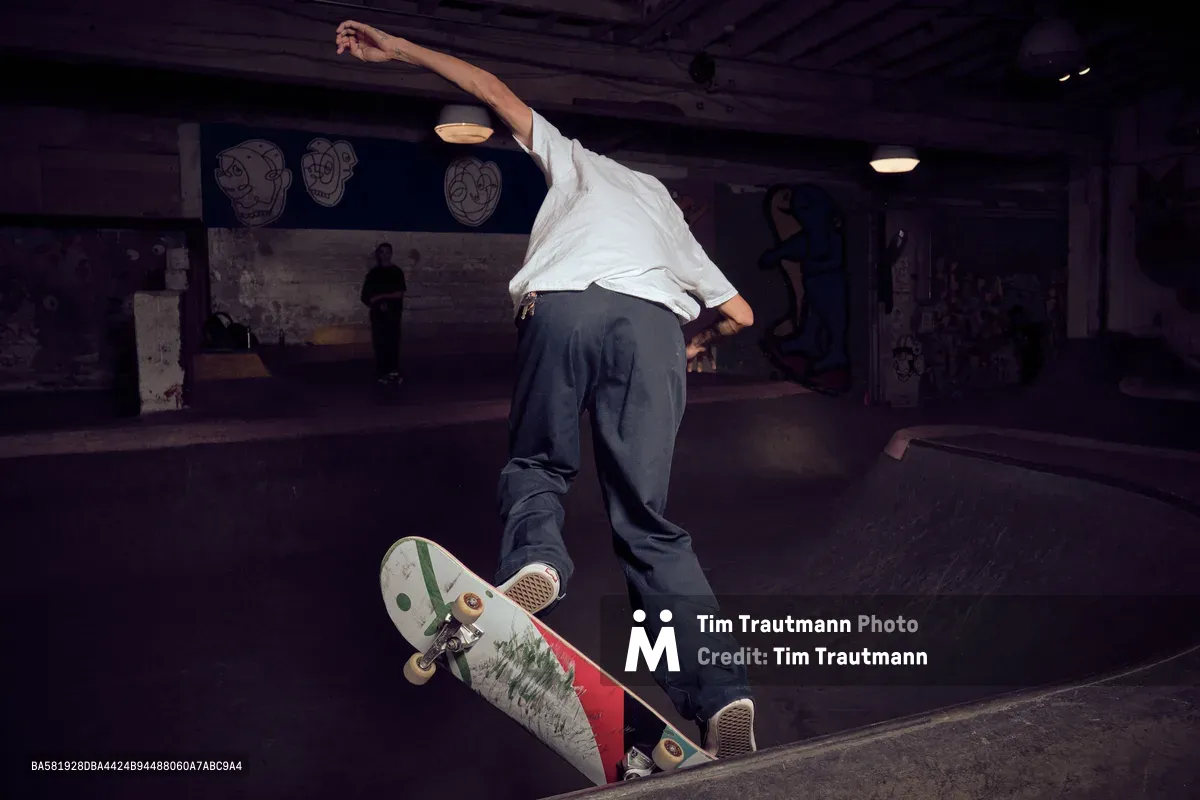 A skateboarder executes a fluid carve through the concrete bowl at Commonwealth Skateboarding in Portland's Buckman neighborhood. The rider, dressed in a crisp white shirt and dark jeans, demonstrates precise board control while navigating the curved transition. Moody tungsten lighting creates dramatic shadows across the weathered concrete surfaces, while fellow skaters observe from the background of this authentic skate sanctuary.
