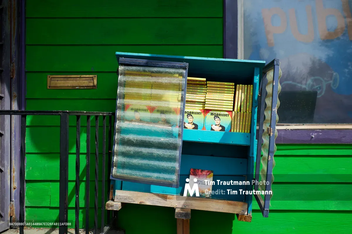 A weathered outdoor book display case sits against the vibrant green clapboard siding of Microcosm Publishing in Portland's Eliot neighborhood. The wooden case with wavy plexiglass doors houses free books including copies of "Defensive Eating with Morrissey," creating a community sharing tableau in the little library tradition. Golden afternoon light illuminates the turquoise-painted display box while a public pay phone stands prominently in the left side of the composition.