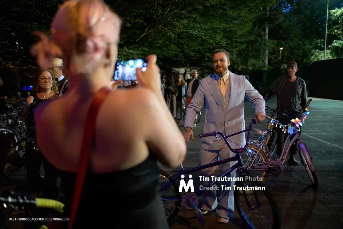 A distinguished man in a pale three-piece suit poses confidently with his decorated bicycle during Portland's whimsical Drop Out Prom Ride at Colonel Summers Community Garden. The evening scene captures the contrast between formal attire and playful rebellion, as smartphone-wielding participants document the unconventional celebration under the canopy of mature trees. Dappled streetlight illuminates the gathering, creating an intimate atmosphere where cycling culture meets tongue-in-cheek formality.