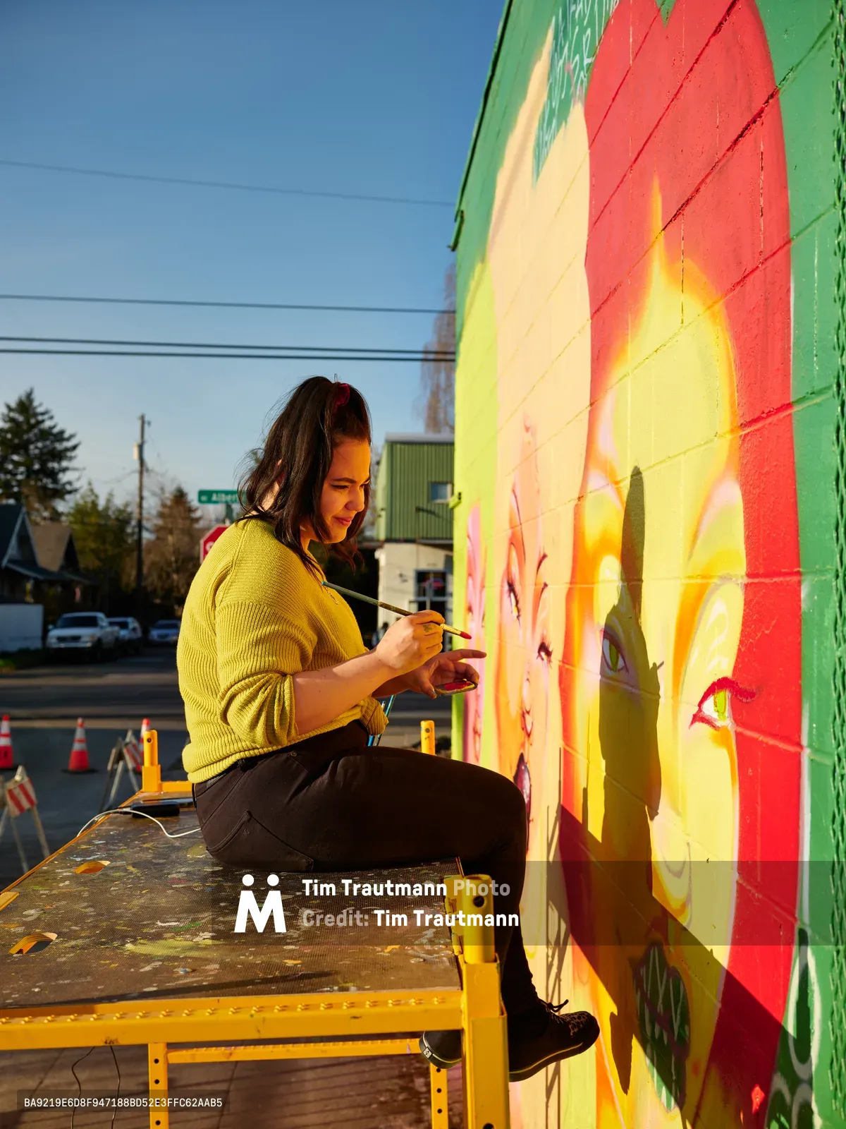 Multidisciplinary artist Vilatose sits cross-legged on a yellow scaffold platform, delicately painting details on her mural titled Jardín Cubana that depicts three female figures on the side of a building in Portland's Alberta Arts District. Golden hour light bathes the scene, casting warm shadows and illuminating her yellow cardigan as she works with focused concentration. The mural features flowing bands of red, orange, yellow, and green pigments that seem to dance across the concrete canvas. Behind her, the quiet residential street stretches toward the horizon under a clear blue sky, dotted with power lines and evergreen trees.