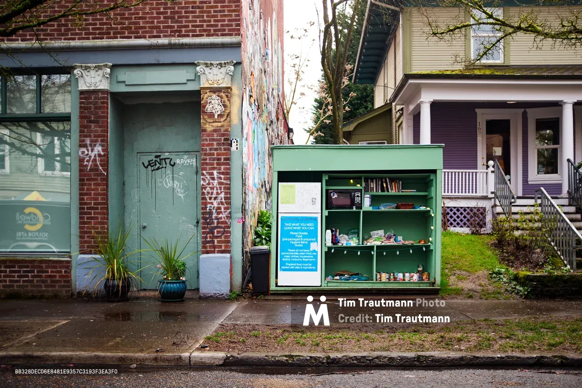 A sage green community refrigerator stands sentinel on Alberta Street in Portland's Vernon neighborhood, its glass-paneled door revealing shelves stocked with donated food and supplies. The weathered brick storefront behind bears graffiti tags while ornate Corinthian columns frame a mint green door, creating a striking juxtaposition between urban decay and grassroots generosity. Purple Victorian houses peek through bare winter branches, capturing the eclectic architectural tapestry of this Northeast Portland corridor where mutual aid meets neighborhood character.