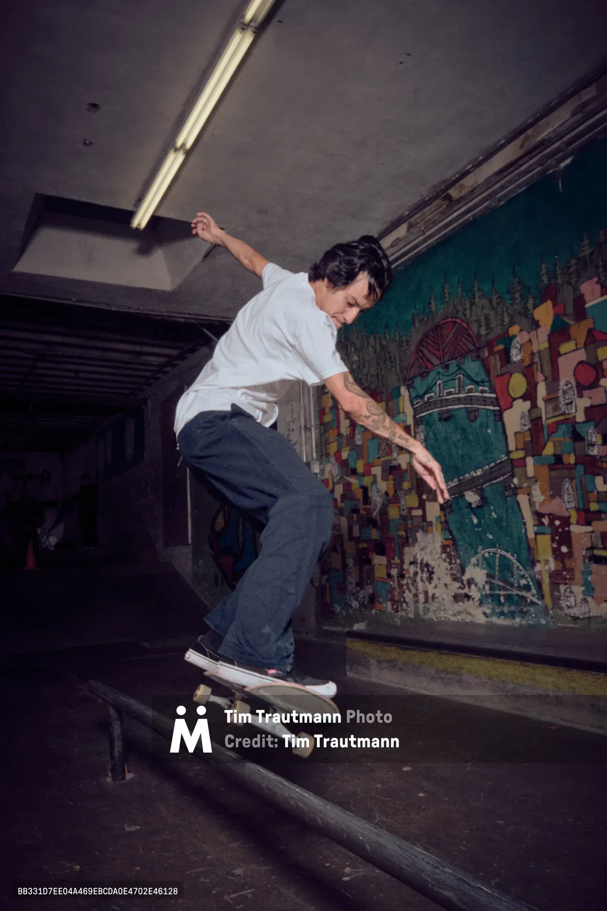 A skateboarder carves through the shadows of Commonwealth Skateboarding's indoor park in Portland's Buckman neighborhood, his white shirt stark against the moody concrete tunnel. Fluorescent lighting cuts harsh angles through the subterranean space while vibrant graffiti murals create a kaleidoscope backdrop of urban art. The rider's fluid motion and focused expression capture the meditative rhythm of street skating in this iconic Pacific Northwest skate sanctuary.