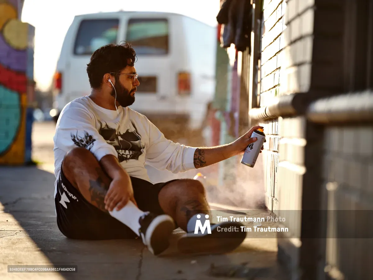 Muralist Heysus sits cross-legged on the sidewalk at Northeast 15th and Killingsworth, spray paint can in hand, bathed in warm golden hour light. The tattooed artist wears earbuds and a graphic tee while working on his latest piece, with colorful graffiti walls and a white van creating an authentic urban backdrop. Long shadows stretch across the concrete as the setting sun illuminates this moment of creative focus in Portland's Alberta Arts District.
